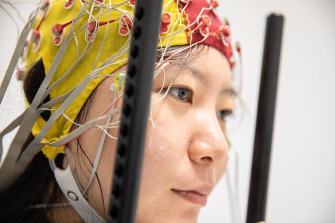 A study participant is facing right, wearing a yellow and red electroencephalogram (EEG) head device that is tight around the participant's head like a swimming cap. The headgear has electrodes attached to it and has a chip strap that goes around the lower part of the participant's face.